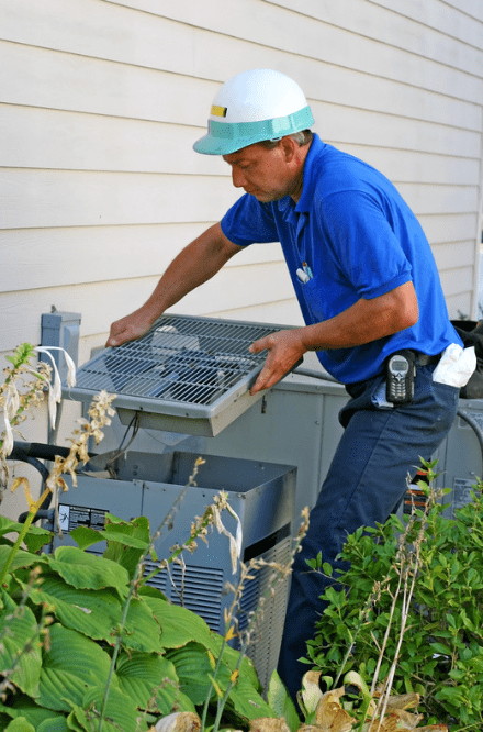 An HVAC specialist in blue pants, blue polo shirt, and a white and blue hard hat removing the top of an outdoor HVAC system to inspect it.